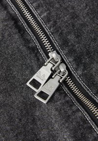 Denim preto com um fecho de duas zipers prateados, apresentando a gravação "Lola's Berlin", realçada por costuras contrastantes nas bordas.