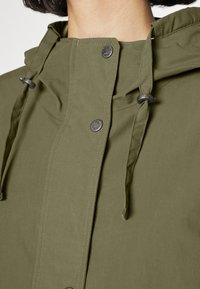 Oliwkowa kurtka z kapturem, zatrzaskami i regulowanymi sznurkami. Miękka tekstura i minimalistyczny design z widocznymi detalami szwów.