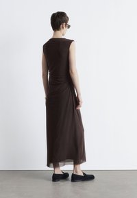 Braunes ärmelloses Kleid mit seitlichem Raffdetail und fließendem Stoff. Kombiniert mit schwarzem Schuhwerk auf einer glatten Oberfläche vor einem weißen Hintergrund.