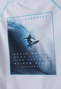 Vit rash guard med en blå grafik av en surfare som rider på en våg, med texten: "SURF PARADISE" och platsinformation i olika stilar.