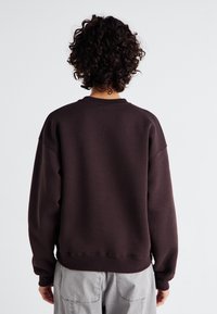 Brun sweatshirt med rund hals och sänkta axlar, med ribbade mudd och nederkant. Slät textur, avslappnad passform, visas bakifrån.