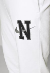 Detalj av vita sweatpants med svart "N" och grå Nike-swooshlogga som överlappar på låret.