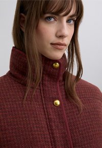 Manteau rouge texturé avec un col haut, arborant des boutons dorés et un design structuré et ajusté. Vue en gros plan mettant en valeur le matériau et les détails.