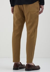 Homme portant un pantalon slim marron et des mocassins en cuir marron foncé, se tenant devant un fond clair et uni.