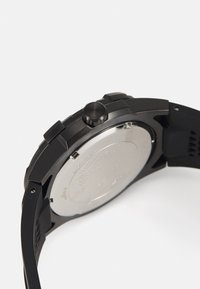 Reloj negro con correa de goma texturizada, caja redonda y parte trasera de acero inoxidable de color plateado. Presenta una corona negra prominente y un diseño elegante.