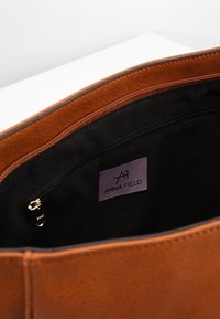 Bruntoff faux läder tote-väska med svart foder, utrustad med en zippered ficka och en etikett märkt "ANNA FIELD."