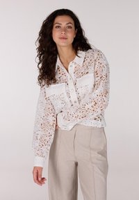 Witte kantblouse met bloempatronen, knoopsluiting aan de voorkant, twee borstzakken en lange mouwen. Draagt met lichte broek.