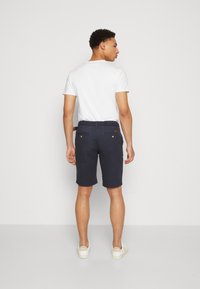 Marineblå shorts med flat front, to baklommer og en vevd midjebånddetalj. Kombinert med en hvit t-skjorte og lyse sko.