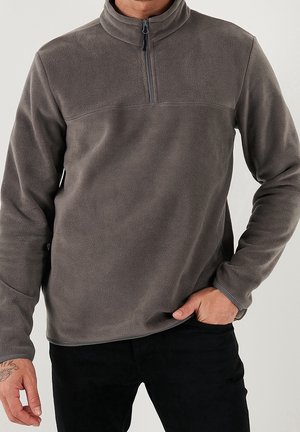 Mężczyzna w szarej polarowej bluzie z krótkim zamkiem, z długimi rękawami, jedną ręką w kieszeni czarnych spodni, widoczne tatuaże na lewym nadgarstku.