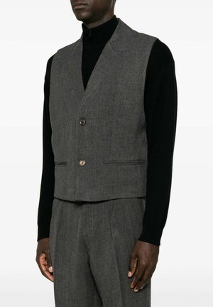 Uomo che indossa un gilet grigio su misura e pantaloni abbinati sopra una maglia nera a maniche lunghe su sfondo bianco.