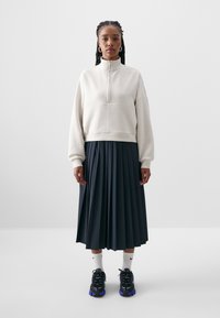 Svetlo bež flis zip-up dukserica v kombinaciji s temno modro plisirano krilo, črnimi supergami in belimi nogavicami. Osredotočite se na sproščen kroj in teksturirane materiale.