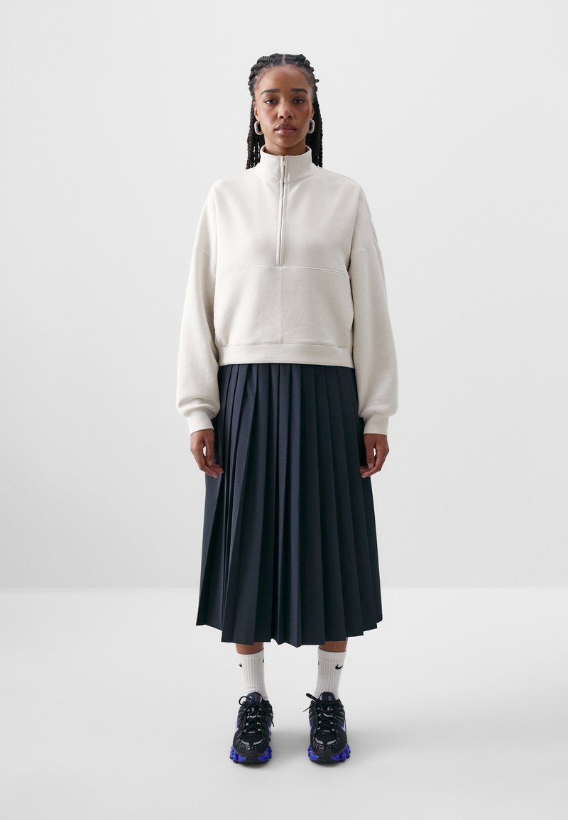 Svijetlo bež majica s poluzipom i opuštenim krojem, uparena s plisiranom tamnoplavom suknjom i crnim tenisicama s ljubičastim detaljima.