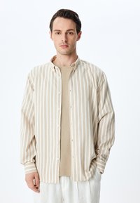 Camisa de botones a rayas beige con un corte relajado, mangas largas y cuello con solapa, superpuesta sobre un suéter beige claro.