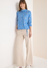 Haut bleu à manches longues avec un motif de feuilles, associé à un pantalon beige à jambes larges ; présente un détail noué à la taille et des baskets blanches.