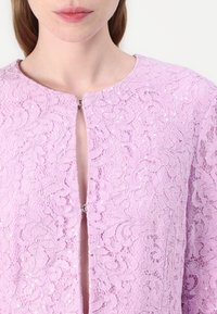 Lila Spitzenkleidung mit komplizierten Blumenmustern, einem runden Ausschnitt und einer zentralen Öffnung, die mit einem silbernen Verschluss gesichert ist. Weiche Textur.