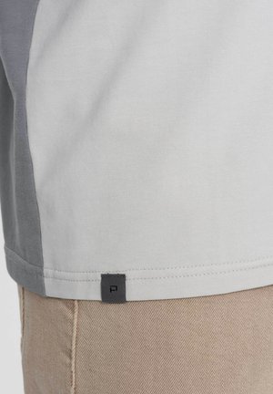 Camiseta bloqueada en colores gris y blanco, con suave tela de algodón, un dobladillo recto y una pequeña etiqueta con el logo gris en la parte inferior izquierda.