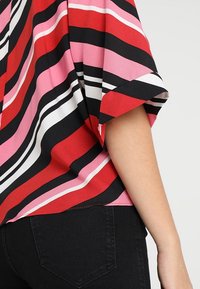 Top a righe multicolori caratterizzato da vivaci tonalità di rosso, rosa, nero e bianco, con una vestibilità comoda e maniche corte. Tessuto morbido e fluido.