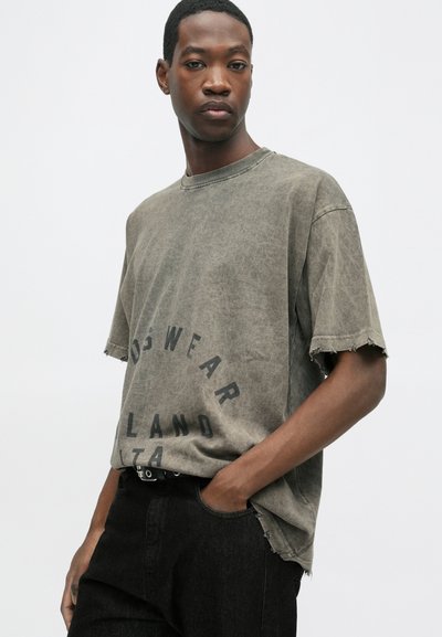 Tricou scurt din țesătură gri închis, cu mâneci scurte și margini ușor destrămate, având text negru imprimat. Purtați-l cu blugi închiși la culoare, evidențiind un design lejer.
