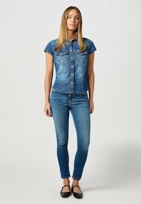 Camisa de denim de manga corta, lavado azul claro, con dos bolsillos en el pecho y cierre de botones, combinada con jeans ajustados azules y bailarinas negras.