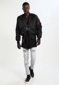 Veste bomber noire avec des poignets côtelés, superposée à une longue chemise noire. Un jean skinny déchiré gris clair et des chaussures noires à enfiler complètent le look.