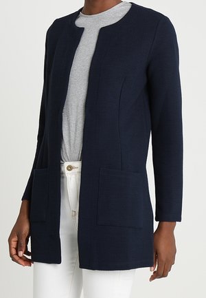 Granatowy kardigan z długim rękawem, otwarty z przodu, z dwoma kieszeniami, noszony na szarej koszuli i białych spodniach.