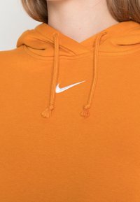Orange hoodie made of textured fabric, featuring a white Nike logo and drawstrings with tassels. Simple and casual design.