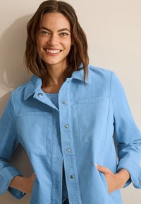 Veste en velours côtelé bleu clair avec une fermeture à bouton, un col pointu, deux poches avant et des manches retroussées. Texture douce et design décontracté.