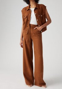 Ruskea denimpaita ilman hihoja rintataskuilla, yhdistettynä korkeavyötäröisiin ruskeisiin leveälahkeisiin housuihin. Sileä rakenne, minimalistikot suunnittelu.