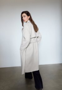 Lang, lys beige frakke med et flydende design, overdimensioneret krave og bælt. Har lommer og en glat tekstur, parret med mørke bukser.