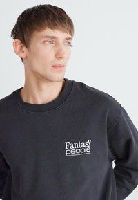 Schwarzer Pullover aus Baumwollgemisch, mit Rundhalsausschnitt und einem weißen Logo "Fantasy people" auf der linken Brust. Einfaches, lässiges Design.