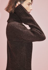 Brauner, gestrickter Rollkragenpullover mit gerippter Textur und langen Ärmeln, mit hohem Kragen und slim fit, vor einem rosa Hintergrund.