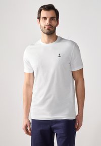 Hvid bomulds t-shirt med korte ærmer, der har et lille sort broderet anker på venstre bryst. Glat tekstur, klassisk pasform.