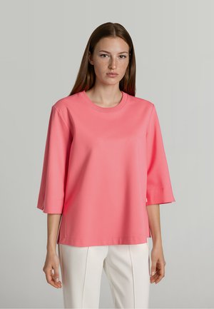 Jonge vrouw met steil bruin haar draagt een losse roze top met driekwart mouwen en witte broek tegen een neutrale achtergrond.