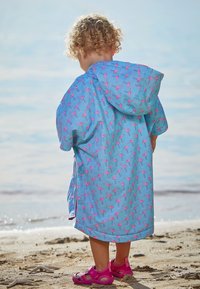 Blå regnponcho med pink flamingo-print, hætte-design og korte ærmer; parret med pink sandaler på en sandstrand.