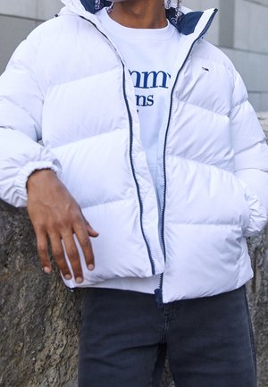 Person iført en hvid puffjakke over en hvid Tommy Jeans-shirt, stående mod en stenmur, hænderne afslappet ned langs siderne.