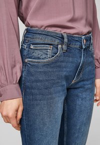 Tmavě modré džínové kalhoty mají střední pas, pět kapes a kontrastní oranžové švy. Hladká textura s mírným efektem vyblednutí.