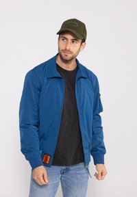Veste bomber bleue avec une fermeture éclair à l'avant, des poignets côtelés et un écusson tissé sur le bas à gauche. Portée sur une chemise noire et accompagnée d'un jean.