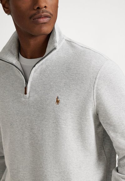 Svetlo siva jopica z zadrgo iz teksturirane tkanine, z majhnim vezenim logotipom konja in visokim ovratnikom s kontrastno zadrgo.