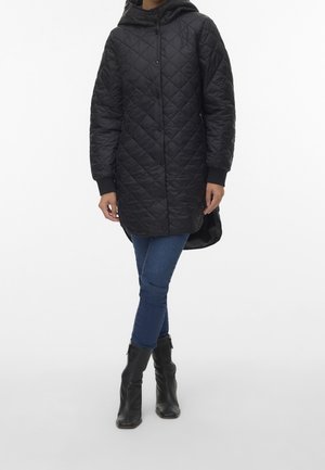 Person iført en sort quiltet hættetrøje, blå skinny jeans og sorte ankelstøvler, stående med krydsede ben mod en ensfarvet baggrund.