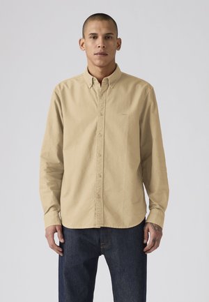 Homme portant une chemise beige boutonnée et un jean foncé, debout devant un fond clair uni.