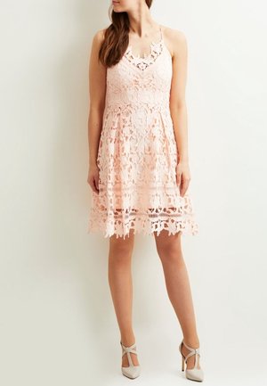 Różowa koronkowa sukienka z kwiatowymi wzorami, dopasowanym gorsetem, regulowanymi cienkimi ramiączkami i rozkloszowanym dołem. Noszona z jasnymi szpilkami.