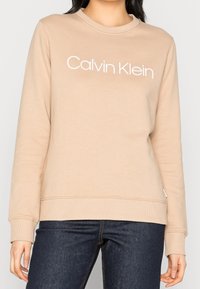 Sudadera beige con cuello redondo, mangas largas y logo blanco de "Calvin Klein" en el pecho. Puños y dobladillo acanalados. Textura de tela suave.