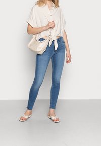 Camisa de botones recortada beis con frente anudado, jeans skinny azules y sandalias de tira crema con diseño anudado, acompañadas de un bolso suave.