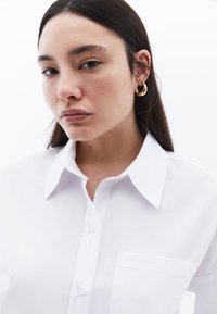 Camicia bianca con bottoni realizzata in tessuto liscio, dotata di colletto, una tasca singola sul lato sinistro del petto e linee pulite.