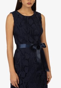 Robe en dentelle marine avec des motifs floraux, sans manches, et un ruban en satin lisse noué à la taille. Détails texturés sur l'ensemble.