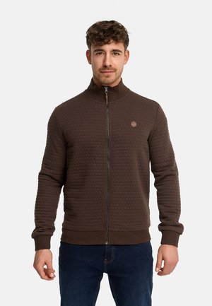 Man draagt een bruin doorgestikt jack met ritssluiting en donkerblauwe spijkerbroek, staand tegen een effen lichte achtergrond, vooruitkijkend.