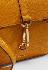 Żółta torebka z ziarnistej skóry ze złotymi okuciami, z zaczepem na klips i tłoczonym logo. Teksturowana powierzchnia i usztywniona konstrukcja.