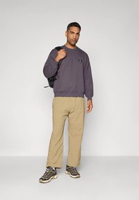 Felpa grigia con piccolo logo, pantaloni beige e scarpe da escursionismo scure con suole chiare. Il modello tiene uno zaino nero.