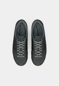 Μαύρα σουέντ αθλητικά παπούτσια με γκρι λεπτομέρειες, με στρογγυλεμένη μύτη, έξι γκρίζα κορδόνια και υφασμένο καουτσούκ πέλμα. Τρεις σκοτεινές πλευρικές ρίγες είναι ορατές.