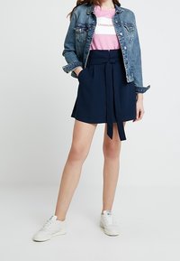 Jeansjacka, rosa t-shirt med vit rand, marinblå paper-bag shorts med knytning, vita sneakers, stående mot en ljus bakgrund.